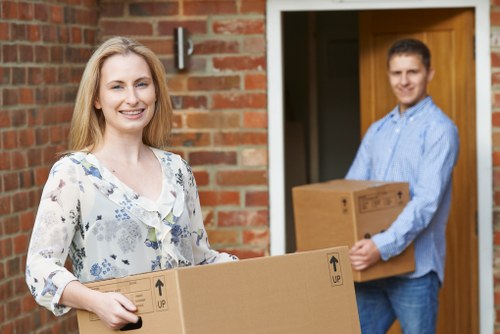 A smiling woman with long blonde hair, wearing a light-colored blouse with floral patterns, is standing outdoors in front of a residential brick house, holding a large cardboard box with black printed arrows indicating the upright position. Behind her, a man with short brown hair, dressed in a light blue shirt and jeans, is standing near an open wooden door, carrying a smaller cardboard box. The background shows the brick exterior of the house and a partially visible garden or outdoor area. The scene suggests a house move, with both individuals engaged in transferring household belongings, including boxes suitable for packing items such as books, decorative objects, or small appliances. The natural daylight highlights the friendly and professional atmosphere of a removals process, consistent with local house moving services in Enfield or nearby areas, as reflected in the context of Enfield Man with Van’s services.