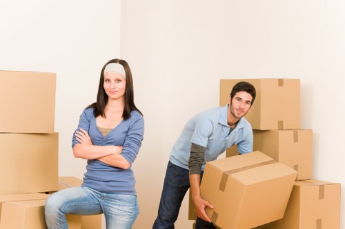 A young woman with long, straight brown hair, wearing a blue long-sleeve top and light blue jeans, is sitting on a cardboard box with her arms crossed and a neutral expression. Beside her, a young man with short dark hair, dressed in a light blue shirt with rolled-up sleeves and dark trousers, is holding a large cardboard box, appearing to be in the process of moving or packing. Several other cardboard boxes of various sizes are arranged around them, some stacked against a plain white wall. The scene is set in an indoor residential space, likely a hallway or a room prepared for a house move. The environment appears clean and well-lit, with natural or artificial light illuminating the scene evenly. The people depicted are engaged in packing or moving activities, consistent with a professional house removals service based in Enfield or nearby areas. Enfield Man with Van offers house removals and related services in the local postcode area.