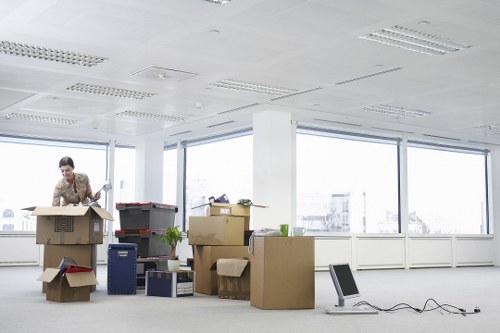 An office scene with a woman in a patterned blazer packing or unpacking items from cardboard boxes on a spacious, well-lit room with large windows showing a cityscape background. The room contains multiple stacked and partially opened boxes of various sizes, some with packing material and others with office supplies or personal belongings visible. A small potted plant sits among the boxes, and a portable computer case is positioned on the floor nearby. The ceiling features recessed lighting, and the floor appears to be covered with a light-colored, professional flooring material suitable for a commercial or residential relocation in Enfield or the surrounding area. The scene captures a moment typical of house or office removals, with the woman actively engaged in organizing or preparing items for transport, reflecting the services offered by Enfield Man with Van, focused on smooth and efficient removals in the local community.