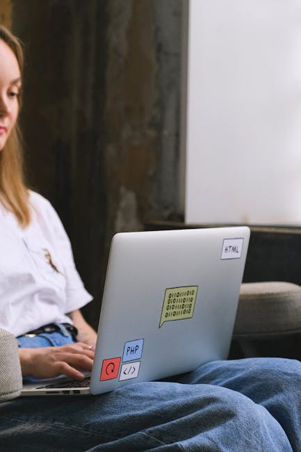 A person typing on a black keyboard in front of a large computer monitor displaying lines of blue and white code or data, situated on a white desk. To the left of the keyboard, there is a white laptop