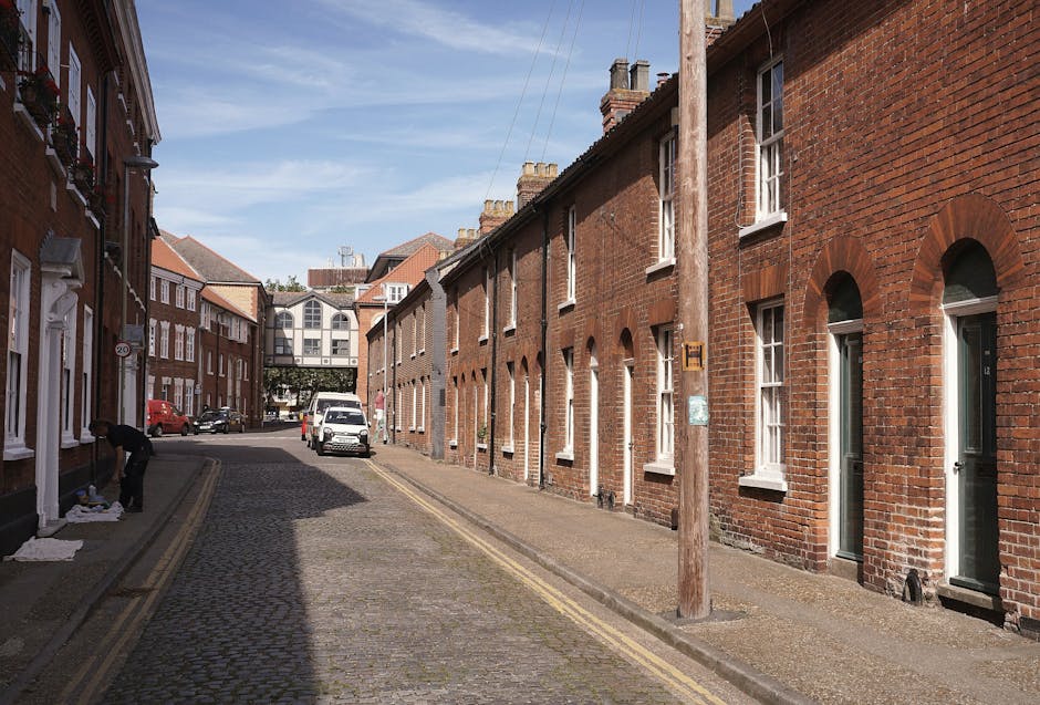 A narrow cobblestone street in an urban residential area with red brick terraced houses on both sides, featuring white-framed sash windows and small doorways. A wooden utility pole stands along the pa